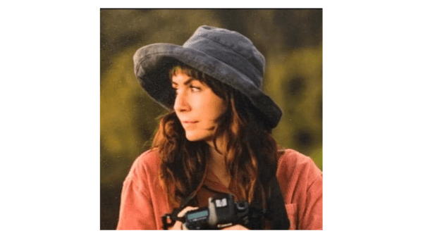 Jessi is standing in a field, wearing a hat and a corduroy shirt, holding a camera and looking to the right. 