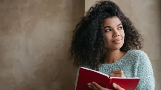 Woman writing in red notebook, looking off to the left.