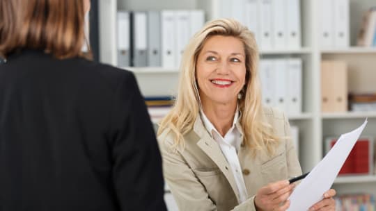 A woman smiles while holding a sheet of paper and looking at another women