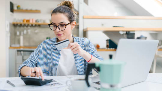 A woman sits at a table while holding a credit card and typing on a calculator