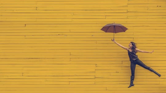 A smiling woman holding an umbrella floats against a yellow background