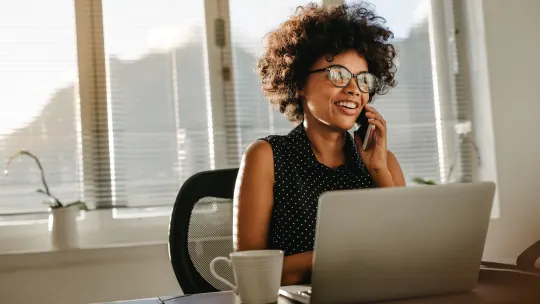 Woman talking on the phone with computer and mug in front of her.