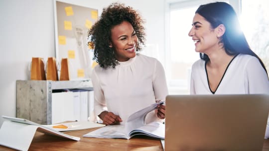 Two coworkers sit at a table and smile while talking