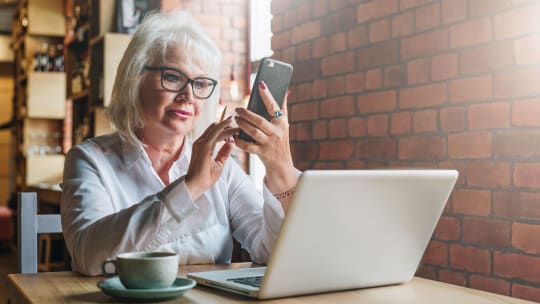 A woman sitting ing from of a laptop looks at her cellphone