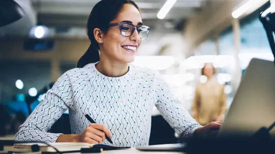 Woman smiling at laptop while taking notes in notebook.
