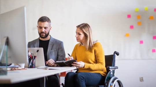 Two coworkers sit in from of a computer and compare notes