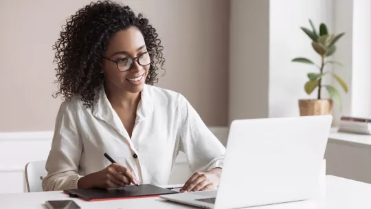 Woman taking notes while smiling at laptop.