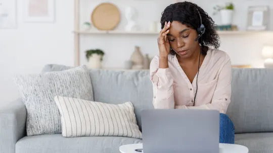 Woman with headset sits on a gray couch with a laptop on a table in front of her. Her right hand is on her forehead and her eyes are closed.