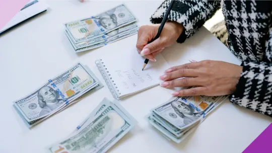 A woman writing calculations in a notebook with four packs of cash on her table to illustrate the importance of learning whether to pay off debt or save money