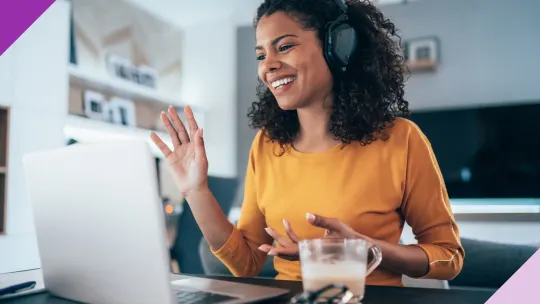 Woman sitting at a desk at home in front of an open laptop wearing headphones and raising one hand in a wave, as though on a video call