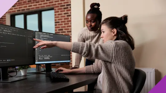 Two women at an office looking at a computer screen showing a coding language to illustrate that it's possible to navigate the gender gap and thrive as women in tech