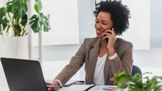 Woman smiles on the phone with laptop and spreadsheets on table in front of her.