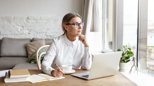 woman staring out of the window on her computer, writing on a notebook