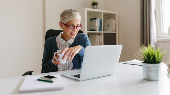 A woman smiles while holding a mug and looking at her laptop