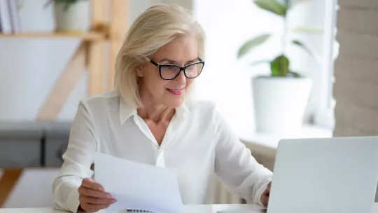 Woman looking at laptop with paper in right hand.