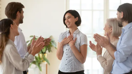 Woman standing, smiling with her hands to her chest with people around her clapping.