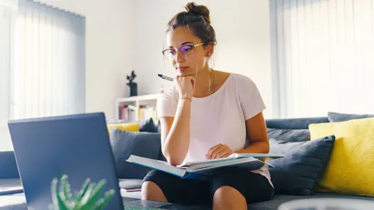 Woman sitting on couch looking at laptop with a notebook in her lap and pen in hand.