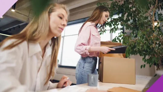 Two women at an office, one is looking at a laptop and one is packing her belongings to leave, illustrating the importance of learning how to emotionally handle being laid off