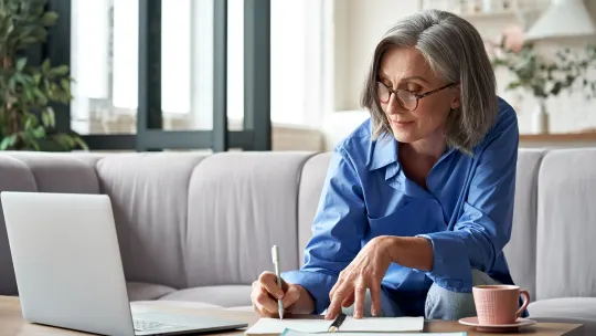 Woman sitting on the couch, writing in a notebook while looking at laptop.