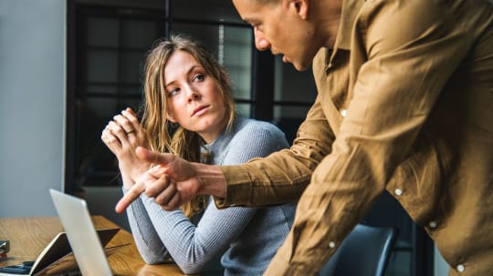 A woman sits at a table while looking at a man explaining something to her