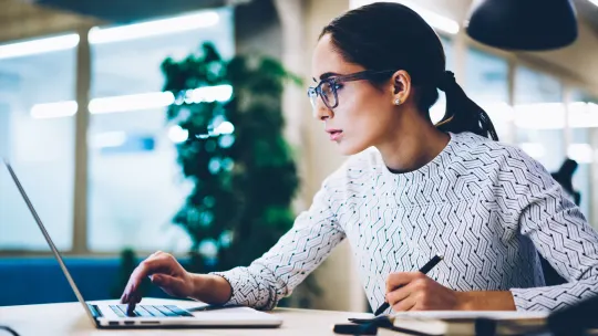Woman sitting at desk looking at computer. She holds a pen in her left hand while taking notes in a notebook.