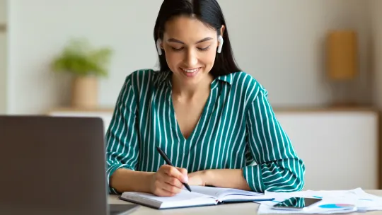 Woman writing notes in notebook with headphones in and laptop in front of her.