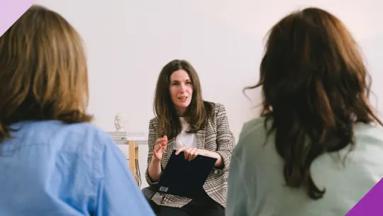 Three women at an office, one of them is teaching something to the others to illustrate why you should learn the difference between coaching vs mentoring