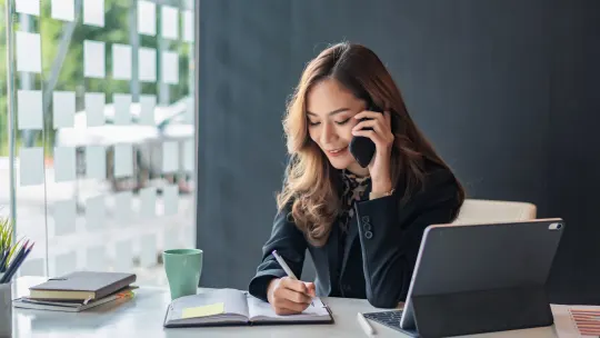 Woman talking on the cell phone while writing in a notebook.
