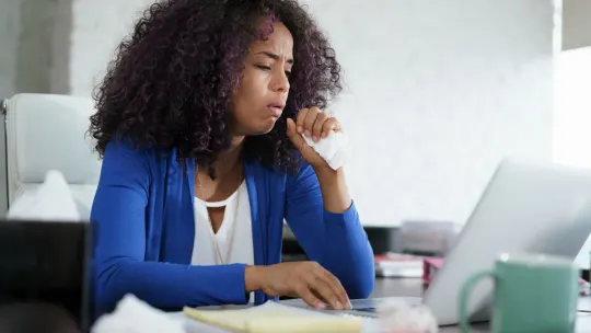 woman working from home coughing into napkin in front of laptop