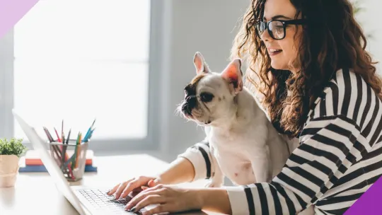 Person sitting at a desk with a dog on their lap, typing on a laptop and smiling