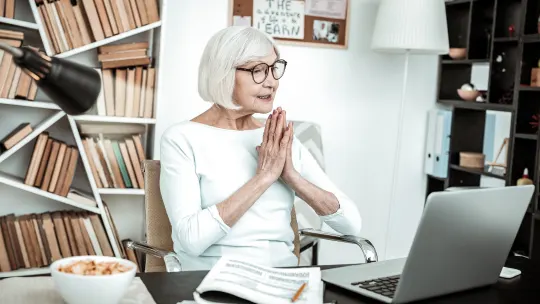 Older woman sitting in front of laptop with hands together in front of her chest.