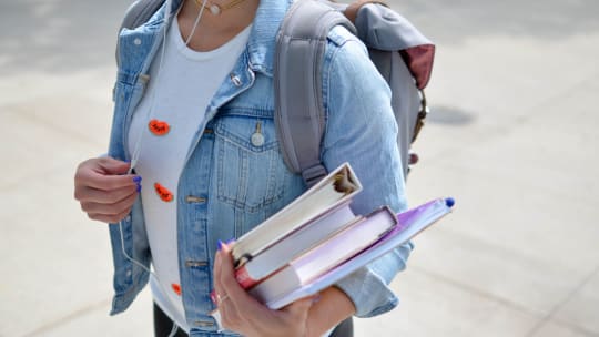 Person wearing a denim jacket and backpack carrying school books 