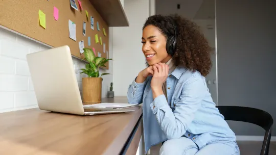 Woman sitting at desk with headphones, smiling at laptop in front of her.