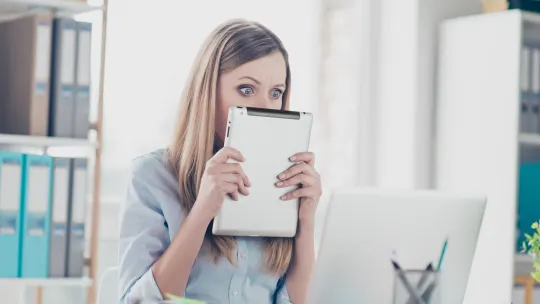 woman covering face with tablet looking nervously at a screen