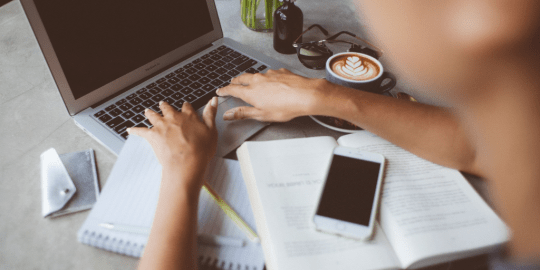 A person typing on a MacBook keyboard with a book and a smartphone next to it