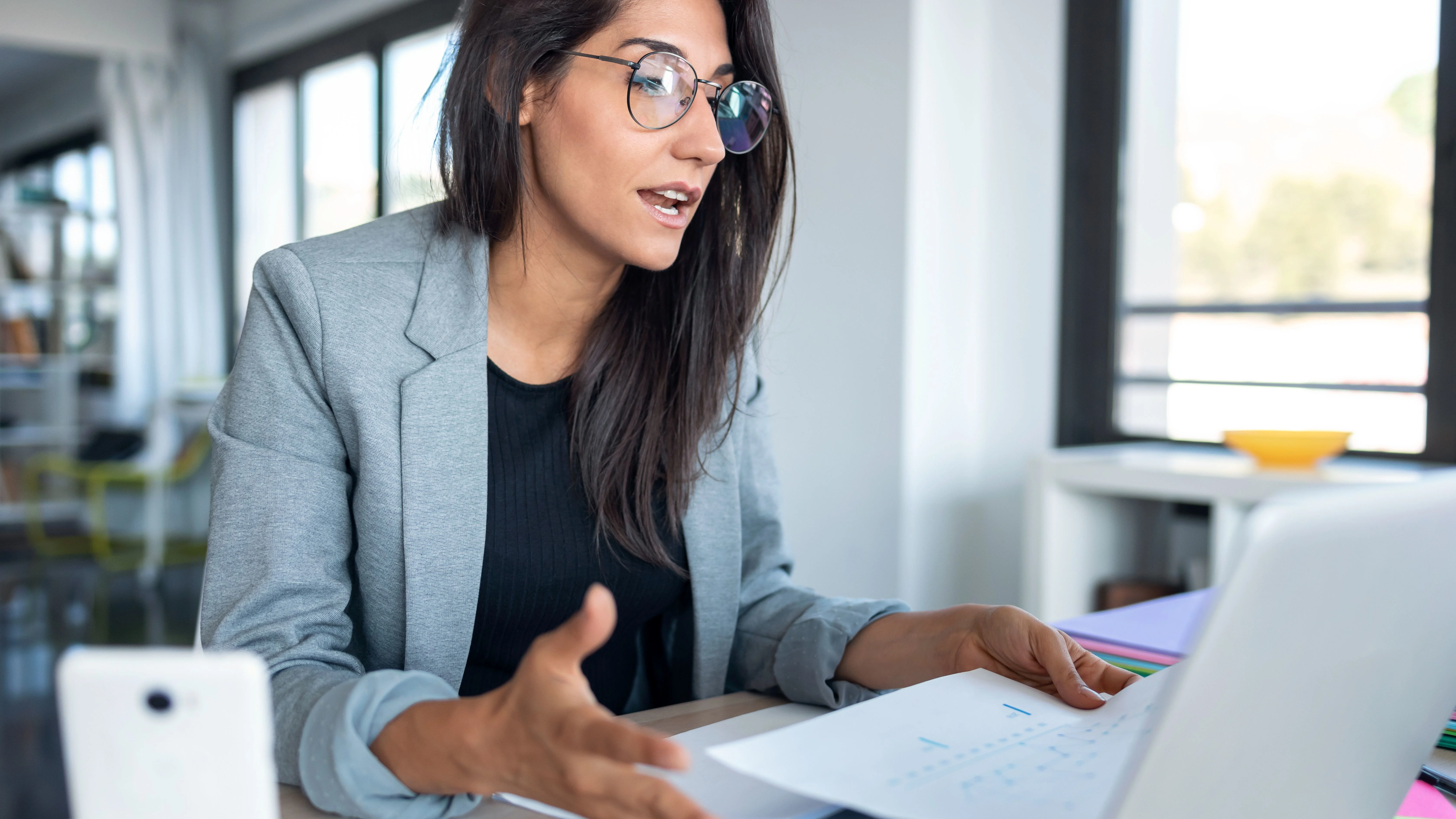 Woman explaining something to laptop with sheet of paper in her left hand.