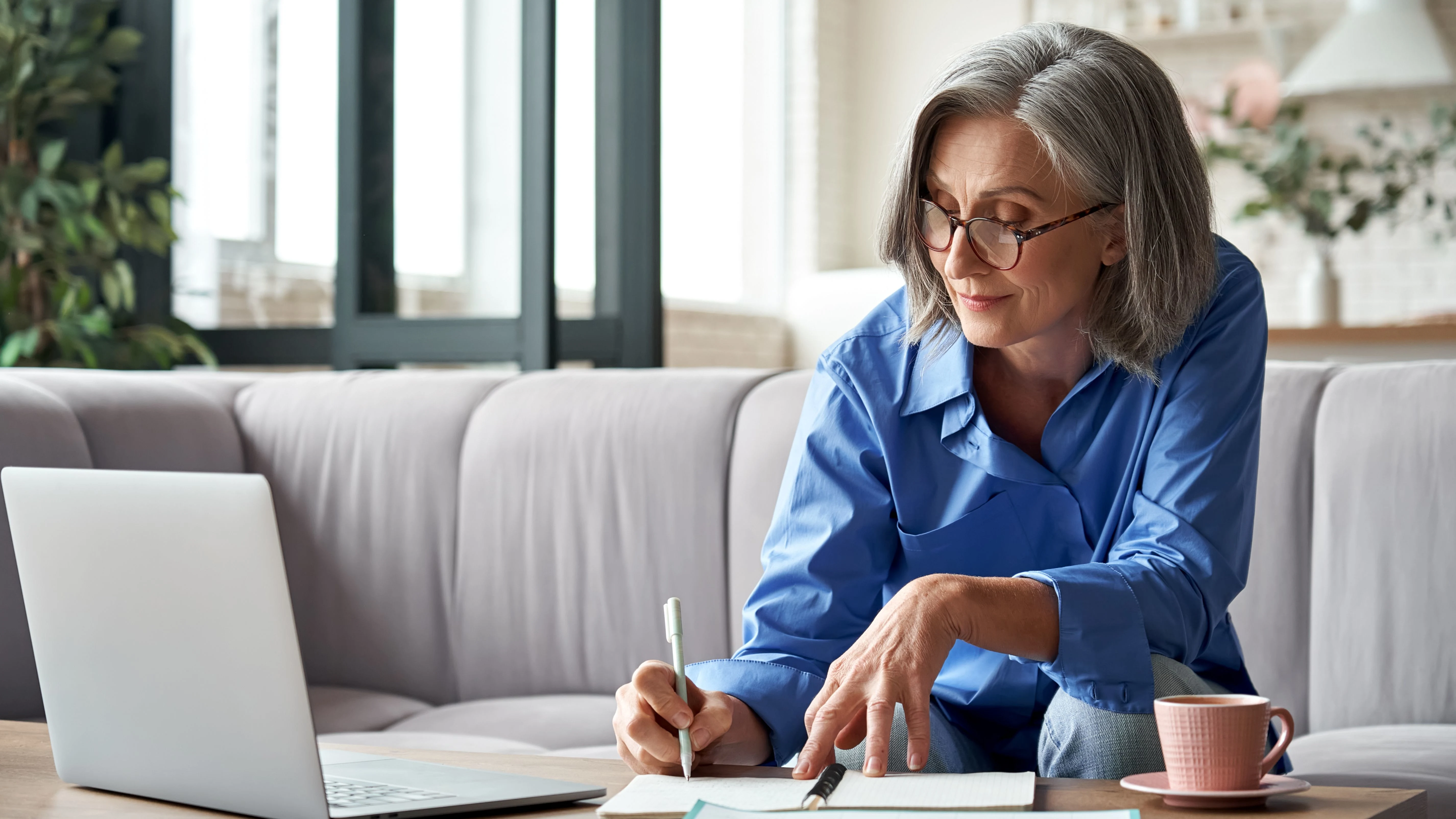 Woman sitting on the couch, writing in a notebook while looking at laptop.