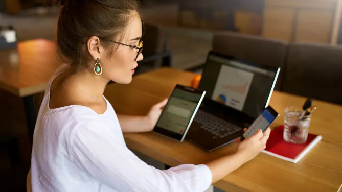 Woman sitting at table looking between her phone, tablet and laptop.