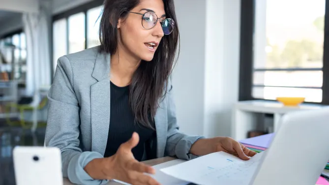 Woman explaining something to laptop with sheet of paper in her left hand.