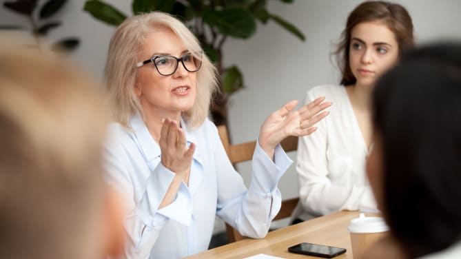 A woman wearing glasses sits at a table surrounded by colleagues