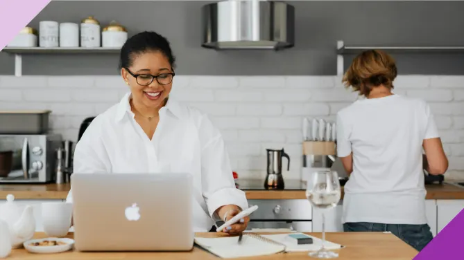 Two women in the kitchen of their house, one of them is managing finances on phone and computer, illustrating how family budget apps can make your life easier