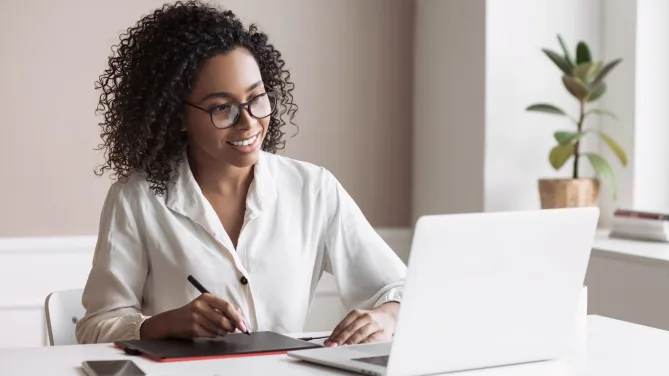 Woman taking notes while smiling at laptop.