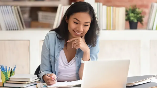 Woman writing in notebook while smiling at laptop in front of her.