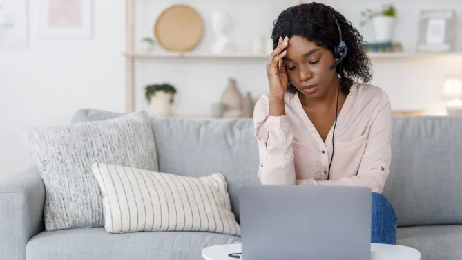 Woman with headset sits on a gray couch with a laptop on a table in front of her. Her right hand is on her forehead and her eyes are closed.