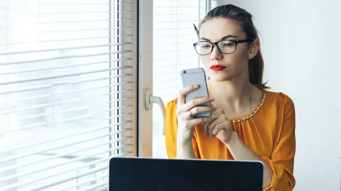 Woman looking at phone with laptop in front of her.