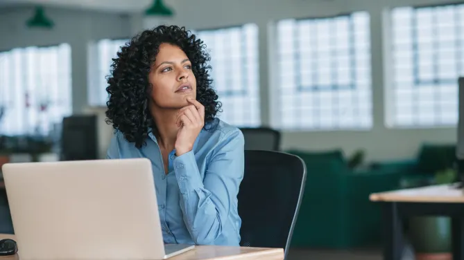 Woman with laptop in front of her looking up right pensively.