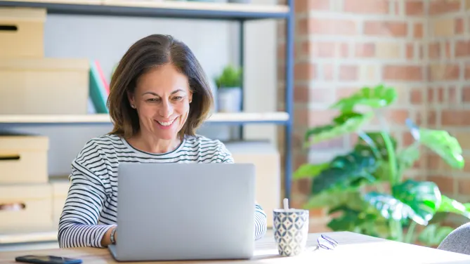 Woman sitting at a table smiling at her laptop with a cellphone, mug, and a pair of glasses on the table.