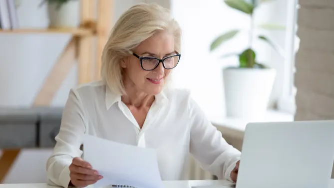 Woman looking at laptop with paper in right hand.
