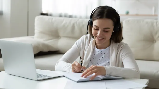 Woman with headset kneeling and writing on notebook on table. Laptop in front of her on the table.