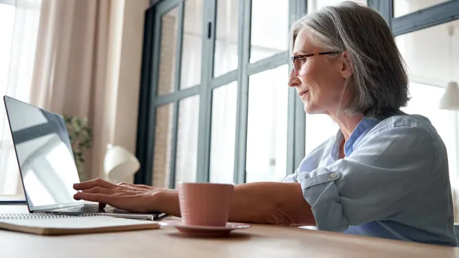 Side view of woman sitting at desk typing on her laptop with a mug beside her.